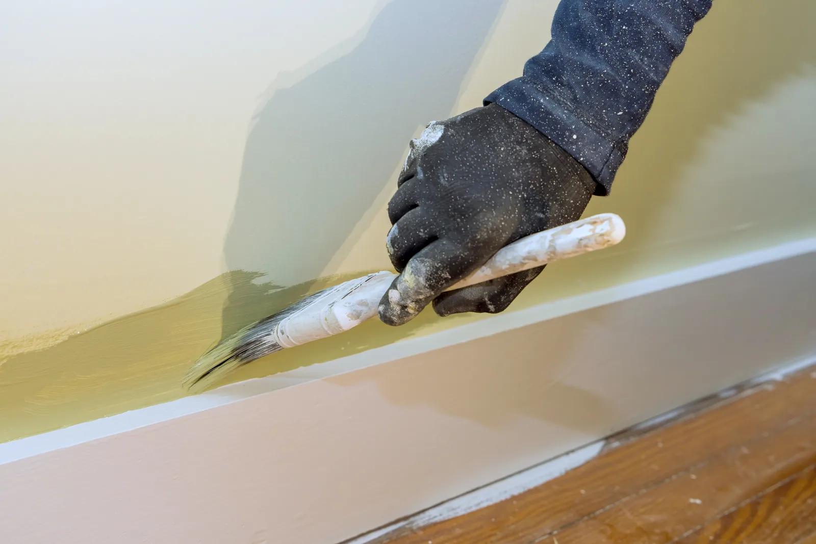 A gloved hand painting a skirting board with a brush, wall masked above
