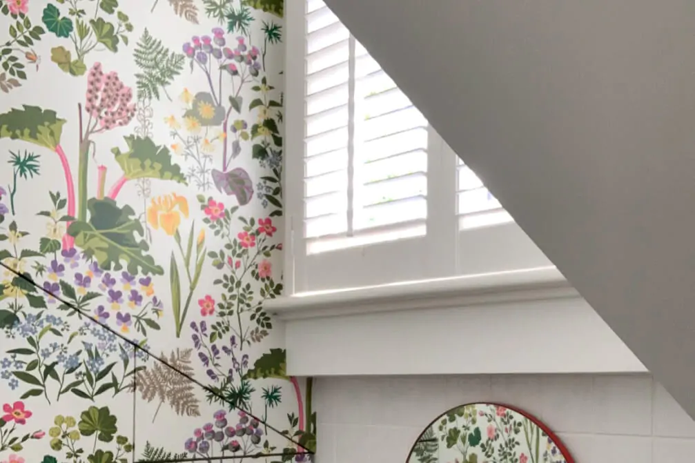 A small bathroom under a staircase features floral wallpaper, a round mirror, and a white shuttered window.