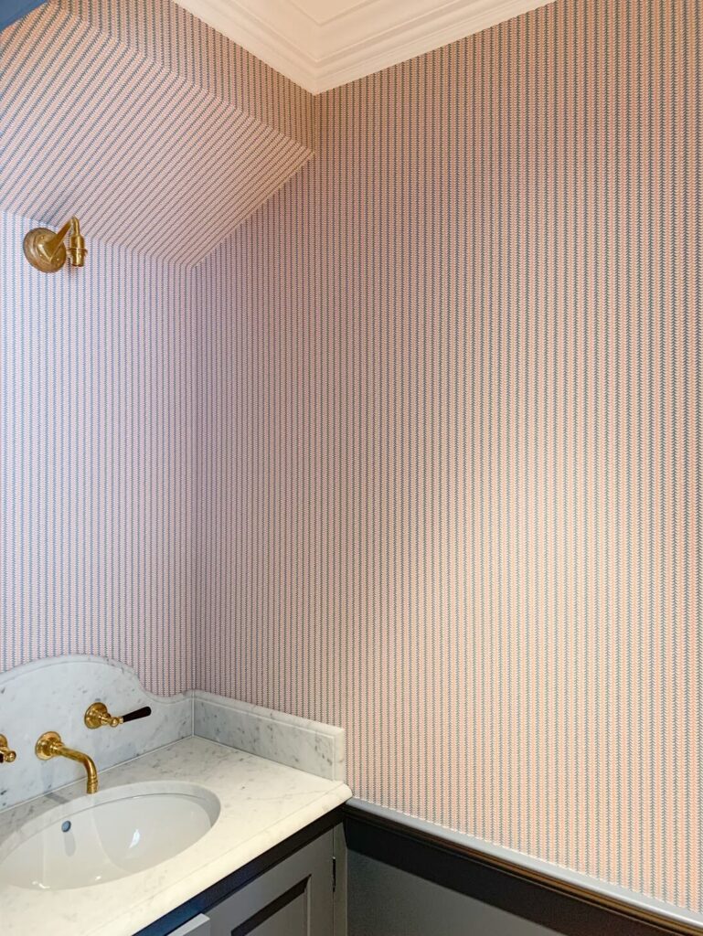 A bathroom corner with pink striped wallpaper expertly installed by wallpaper hangers in London, featuring a gold wall-mounted light, a marble countertop with an undermount sink, and elegant gold faucet fixtures.