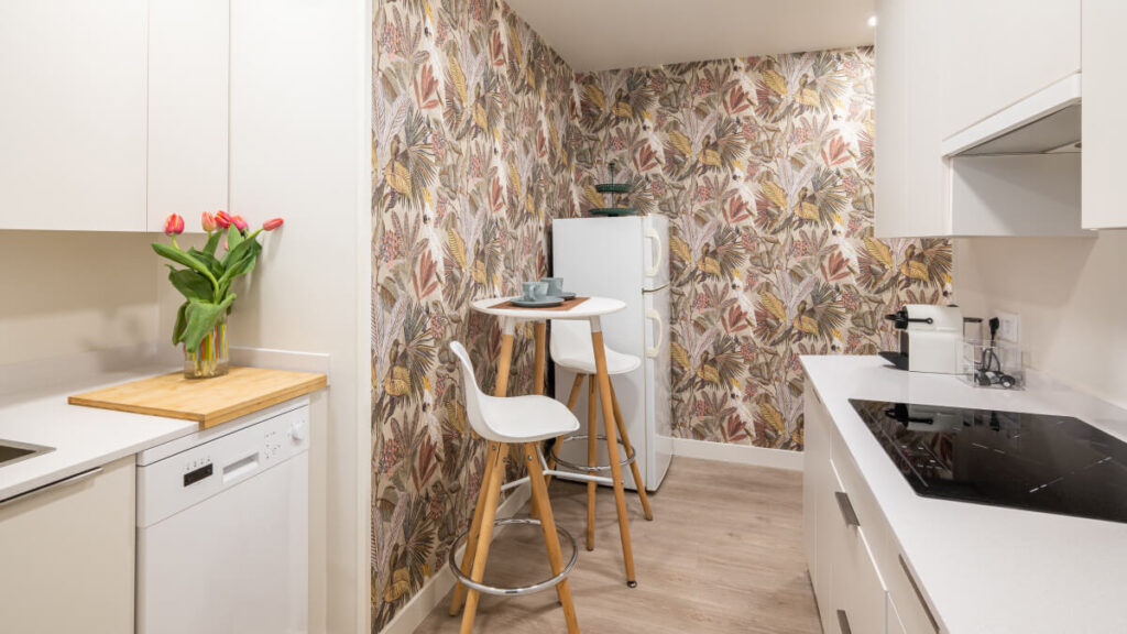 Compact kitchen with floral wallpaper, a small round table with two white stools, white cabinets, a fridge, stove, dishwasher, and a vase of red tulips on a cutting board.