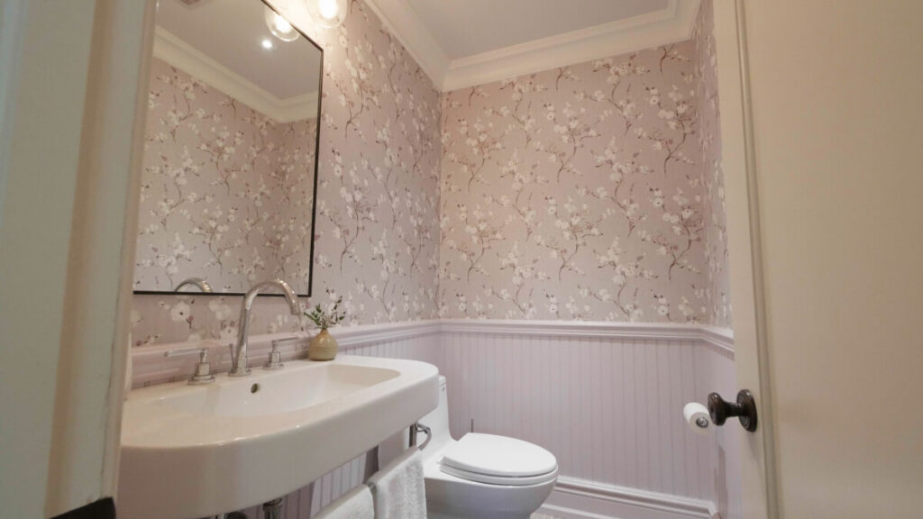 Small bathroom with floral wallpaper, a white pedestal sink, wall-mounted mirror, toilet, and light fixture above. White wainscoting and a towel hanging below the sink.