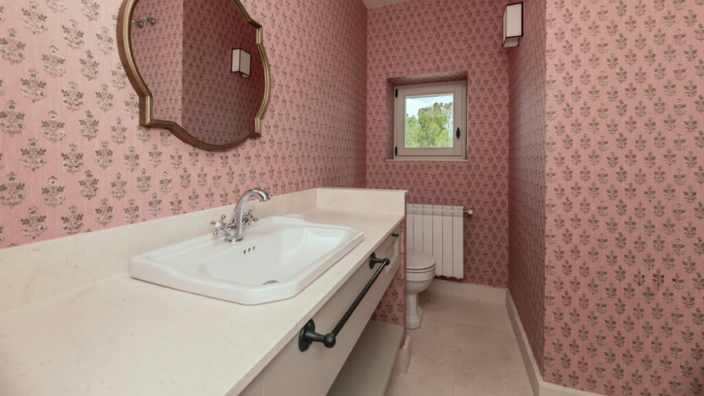 A bathroom with pink floral wallpaper, a white sink with a marble countertop, a wall mirror, a toilet, a window, and a wall light fixture.