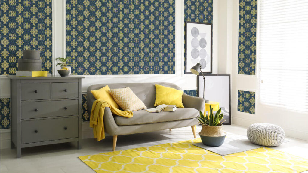 A living room with a gray sofa, yellow and gray cushions, a gray dresser, wallpaper in wet rooms-inspired blue and yellow patterns, a yellow rug, plants, wall art, and large windows with blinds.