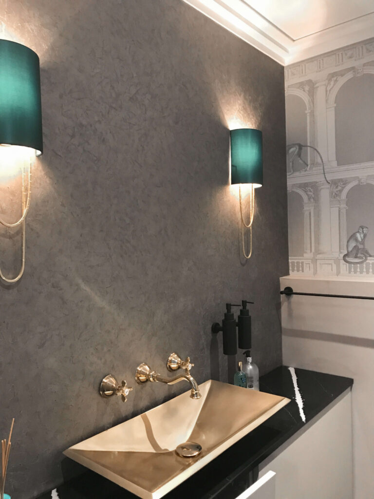 A modern bathroom with a gold geometric sink, black countertop, wall-mounted soap dispensers, two green wall sconces, and Wallpaper in Wet Rooms featuring a classical gray illustration.