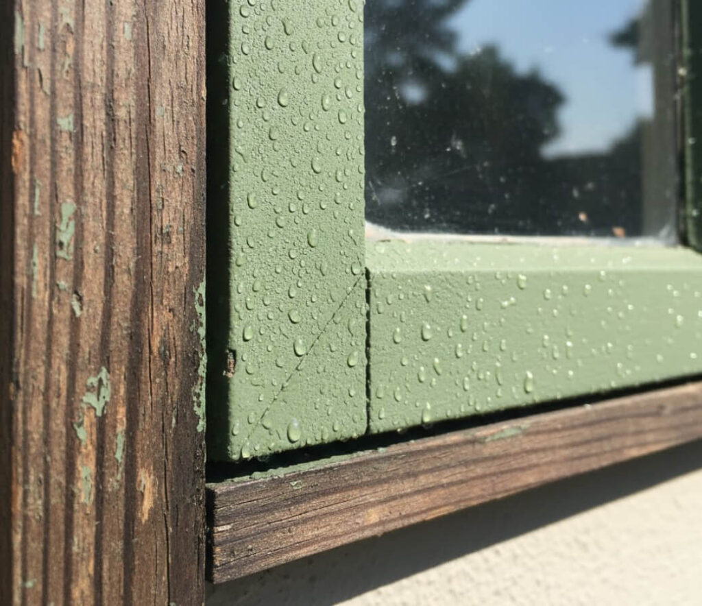 Close-up of a weathered sash window restoration, featuring a green-painted wooden frame with water droplets. Sunlight creates striking shadows and highlights on the textured wood and aged paint.