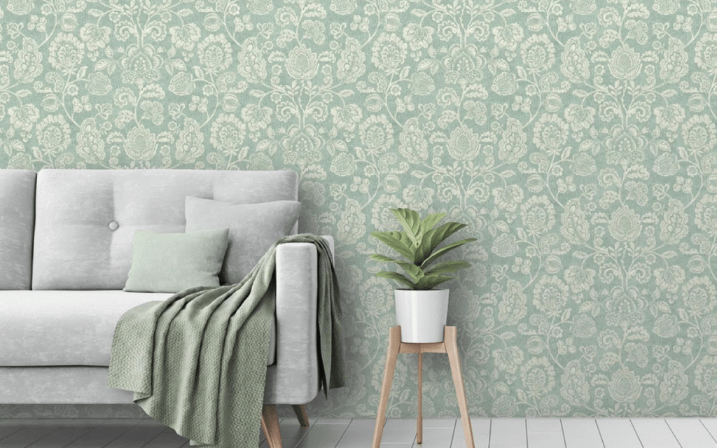 A light gray sofa with a green pillow and blanket stands next to a potted plant on a wooden stand, set against green floral-patterned wallpaper—a stylish nod to current Wallpaper Trends.