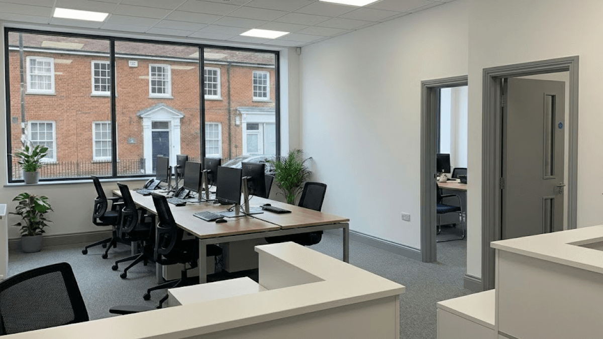 Modern office with several desks, office chairs, and computer monitors near a large window; outside, a street lined with brick buildings featuring Winter-Ready Exteriors is visible.