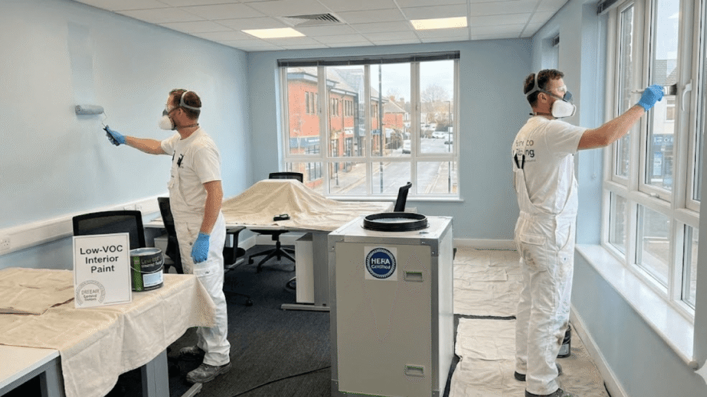 Two painters wearing masks and gloves apply paint to the walls of a modern office, with furniture covered, "Low-VOC Interior Paint," a HEPA air purifier, and tips for keeping Winter-Ready Exteriors visible in the workspace.