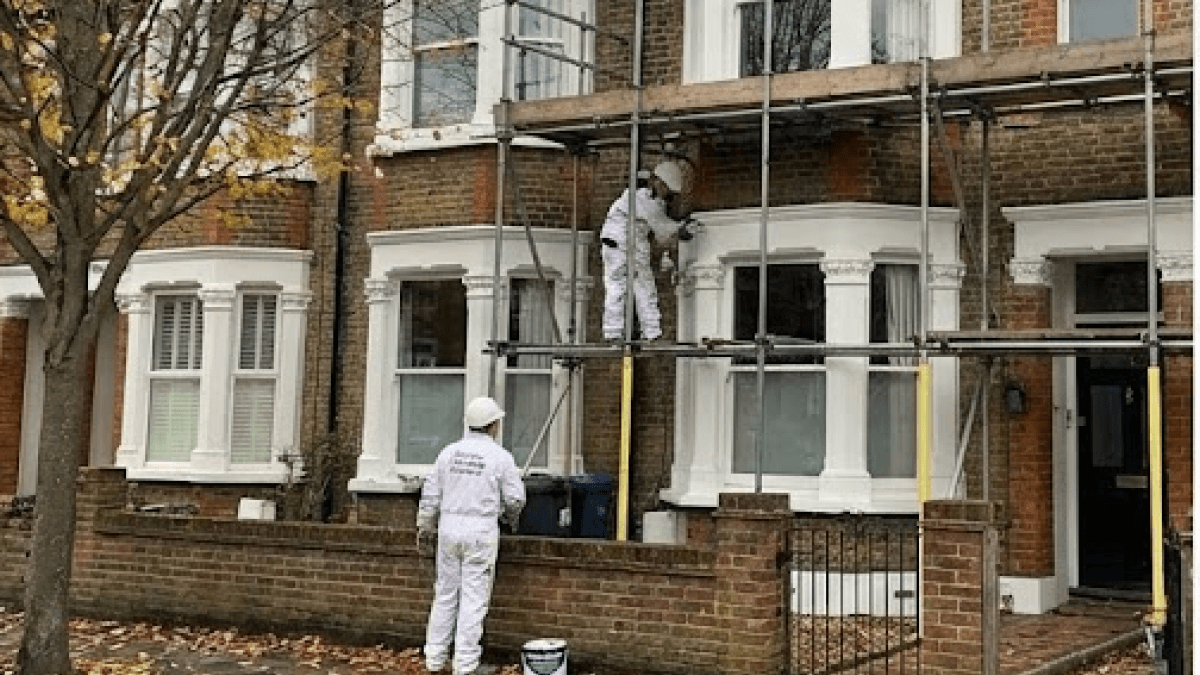 Two workers in white protective suits are painting or renovating the exterior of a brick house on scaffolding, ensuring Winter-Ready Exteriors for lasting protection.