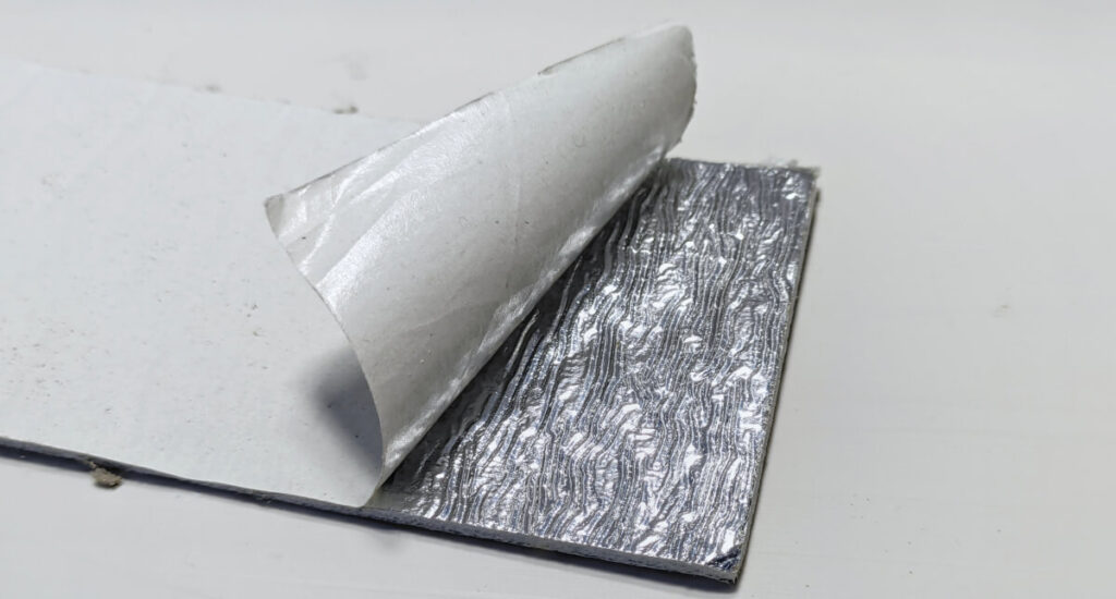 A sheet of material with a metallic, textured adhesive layer—ideal for pairing with thermal paint in Surrey to lower heating bills—is partially peeled back from its backing paper on a flat surface.